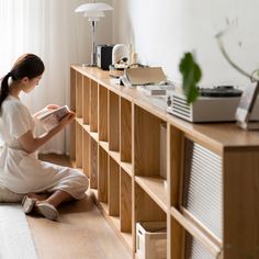 a woman sitting on the floor reading a book in front of a wooden shelf with books