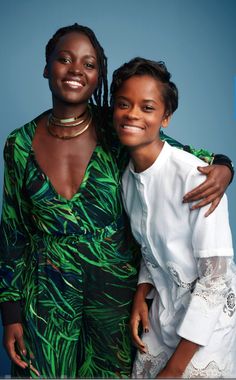 two women standing next to each other in front of a blue background, one is wearing a green dress and the other wears a white shirt