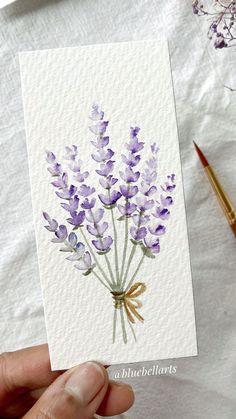 a hand holding a card with watercolor lavender flowers on it and a brush next to it