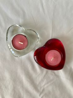 two red candles in a heart shaped glass bowl on a white bed sheet, one is lit and the other is empty