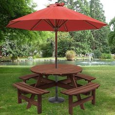 a picnic table with two benches under an umbrella in the grass near a pond and trees