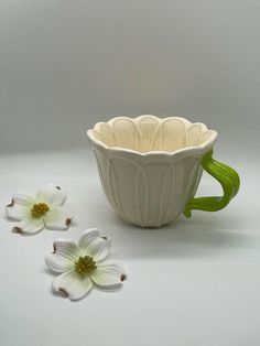 two white flowers are sitting next to a ceramic coffee cup with green handles and handle