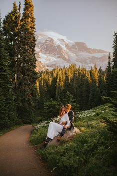 two people sitting on a rock in the middle of a forest with mountains in the background