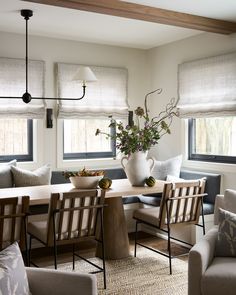 a dining room table surrounded by chairs and vase with flowers on it in front of two windows