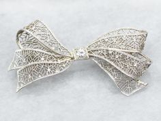 Add a touch of elegance and whimsy to any outfit with this bow brooch. The delicate filigree detailing and sparkling European Cut diamond make this brooch a timeless statement piece. Perfect for any occasion! Metal: 14K White Gold Gem: European Cut Diamond .10 Carats, SI1 in Clarity, I in Color Gem Measurements: 3.2 mm, Round Measurements: 44 x 23 mm Marks: "X14K" Stamped on the pin guard SKU #: A39573 Each piece has been identified and graded by a Graduate Gemologist who has been certified by the Gemological Institute of America (GIA). We have six brick-and-mortar storefronts in Maine, Massachusetts, and New Hampshire and have been in business for over 25 years! Please visit our Shop's About Page or our website for more information about our jewelry. For questions about diamond grading, w White Gold Brooches With Intricate Design For Wedding, Wedding Brooch With Intricate Diamond Design, White Gold Wedding Brooches With Intricate Design, Elegant Filigree Brooches For Evening, Wedding Diamond Brooch With Intricate Design, Diamond Filigree Brooches As Gift, Elegant Brooches With Intricate Design For Anniversary, Elegant White Gold Brooch With Intricate Design, Elegant White Gold Brooches With Intricate Design