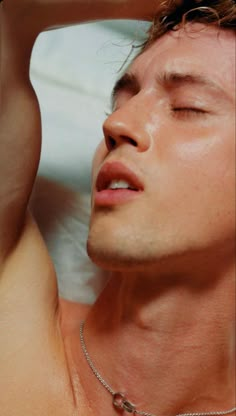 a shirtless man with his eyes closed laying on a white sheet and wearing a silver necklace