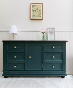 a green dresser with gold handles and drawers in a white walled room next to a painting on the wall