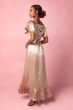 a woman in a gold dress standing against a pink background with her back to the camera