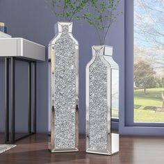 two silver vases with flowers in them sitting on a wooden floor next to a window