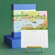 a desk calendar sitting on top of a blue stand next to a card holder with an image of a boat in a pond