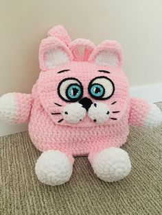 a crocheted pink cat with big eyes sitting on the floor next to a wall