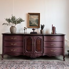 an antique dresser with two vases on top of it next to a painting and potted plants