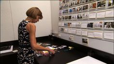 a woman standing in front of a table with magazines on the wall and photos hanging up behind it