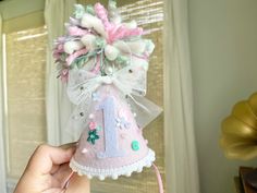 a hand holding a small pink and white birthday hat with flowers on the top, in front of a window