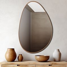 an oval mirror hanging on the wall above a dresser with vases and bowls in front of it