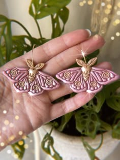 two pink and gold butterfly shaped earrings on a person's hand next to a potted plant