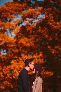 a man and woman standing next to each other in front of trees with orange leaves