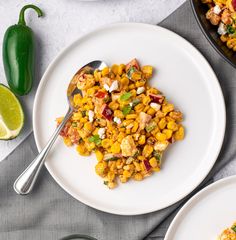 two plates with mexican corn salad on them next to lime wedges and jalapenos