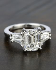 an emerald cut diamond ring with three baguets on the side, sitting on a table