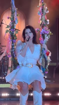 a woman in white dress and boots on stage with flowers hanging from the ceiling behind her