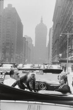 three people in a convertible car on a city street with tall buildings and skyscrapers