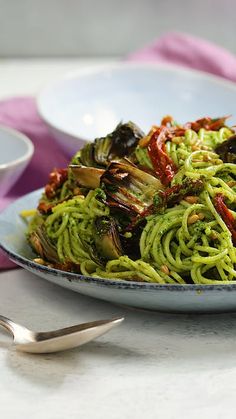 a plate full of green pasta and vegetables on a table next to two silver spoons