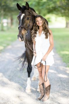 a beautiful young woman standing next to a brown and white horse on a dirt road