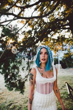 a woman with blue hair and tattoos standing in front of a tree wearing a white dress