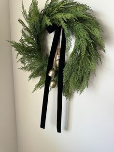 a green wreath with bells hanging on the wall next to a white wall and black ribbon