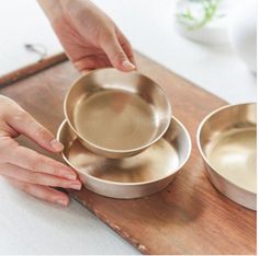 two hands are reaching for three bowls on a wooden tray, one is silver and the other is gold