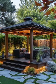 A cozy, well-lit outdoor gazebo with cushioned chairs and plants, surrounded by lush greenery and stone walkways, creating a relaxing backyard retreat. Raised Gazebo Ideas, Home Gazebo, Gazebo In Garden, Sit Out Design, Gazebo Roof Ideas, Gazebo Design Outdoor, Luxury Gazebo, Backyard Gazebo Ideas, Gazebo Ideas Backyard