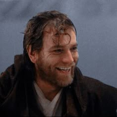 a man with wet hair and beard smiles at the camera while wearing a brown jacket