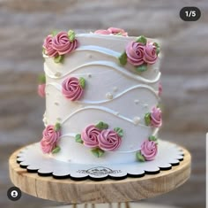 a white cake with pink roses on it sitting on top of a wooden stand next to a brick wall