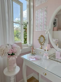 a white desk topped with a mirror and a vase filled with pink flowers next to a window