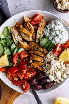 a white bowl filled with chicken, cucumber, tomatoes and other food items