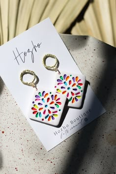 a pair of earrings with colorful designs on them sitting on top of a table next to a card