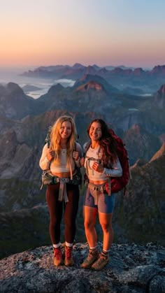 two young women standing on top of a mountain at sunset with mountains in the background