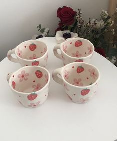 four cups with strawberry designs on them sitting on a table next to flowers and a vase