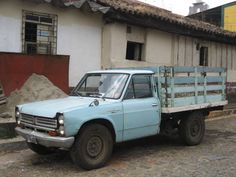 an old blue pick up truck parked in front of a white and gray building on a cobblestone street