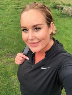 a woman holding a cell phone in her right hand and smiling at the camera with grass behind her