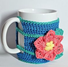 a crocheted coffee cup holder with a flower on the front and bottom, sitting next to a white mug