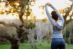 a woman is stretching her arms in the air while standing on a grassy area with trees behind her