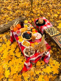 a picnic table set up in the woods with fall leaves on the ground and food laid out on it