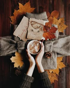 two hands holding a cup of hot chocolate in front of autumn leaves and other items