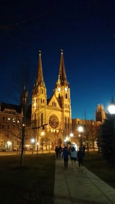 people are walking down the sidewalk in front of a cathedral at night time with lights on