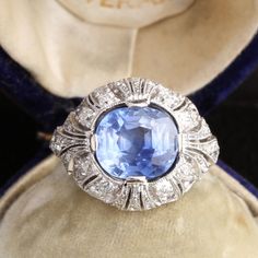 This exquisite ring was fashioned at the height of the Edwardian period. The masterfully crafted mounting is made in platinum and contains wonderfully rendered decoration: intricate engraving, filigree work, feminine ribbon motifs, a generous sprinkling of fine milgrain and 1.10ctw of old mine cut diamonds. A regal 4.25ct cushion cut periwinkle blue sapphire dazzles in the ring's central setting. This richly saturated, beautifully faceted blue stone hails from Sri Lanka (not so long ago known Platinum Diamond Cut Rings For Evening, Elegant Formal Topaz Ring With 17 Jewels, Elegant Platinum Topaz Ring With Brilliant Cut, Evening Platinum Rings With Brilliant Cut, Platinum Rings With Brilliant Cut For Evening, Art Deco Platinum Sapphire Ring With 17 Jewels, Art Deco Platinum Sapphire Ring, Art Deco Sapphire Ring With Platinum Center Stone, Collectible Art Deco Platinum Sapphire Ring