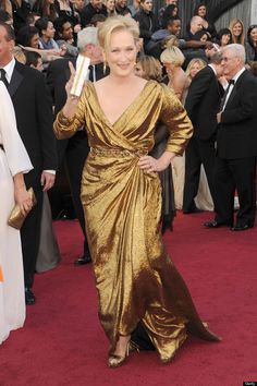 a woman in a gold dress waves to the crowd at an oscars red carpet event