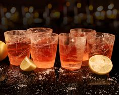 four glasses filled with ice and lemon on top of a black table next to two lemon wedges