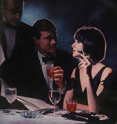 a man and woman sitting at a table with wine glasses in front of them, looking at each other