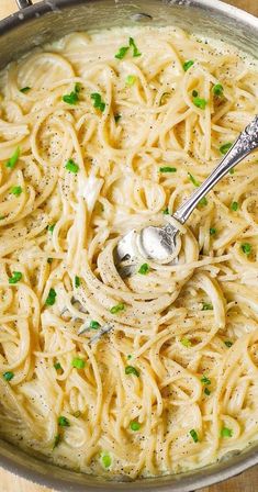 a large pot filled with pasta and parsley on top of a wooden table next to a silver spoon